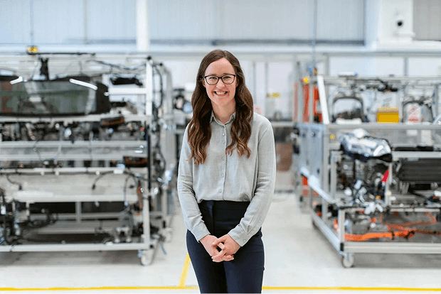 A woman wearing glasses standing in an industrial facility with shelves of mechanical parts in the background